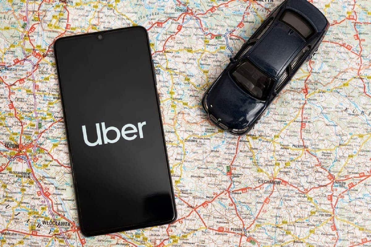Imagem de um celular com o aplicativo da Uber aberto. Ao lado, um carro preto. Ambos estão sobre um mapa.