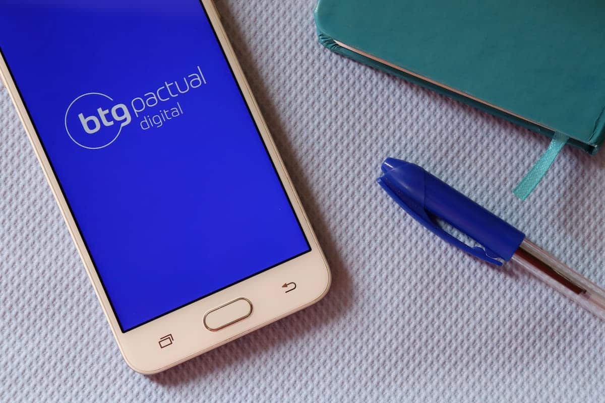 Aplicativo BTG Banking do BTG Pactual em celular de cliente sobre mesa ao lado de caderno e caneta