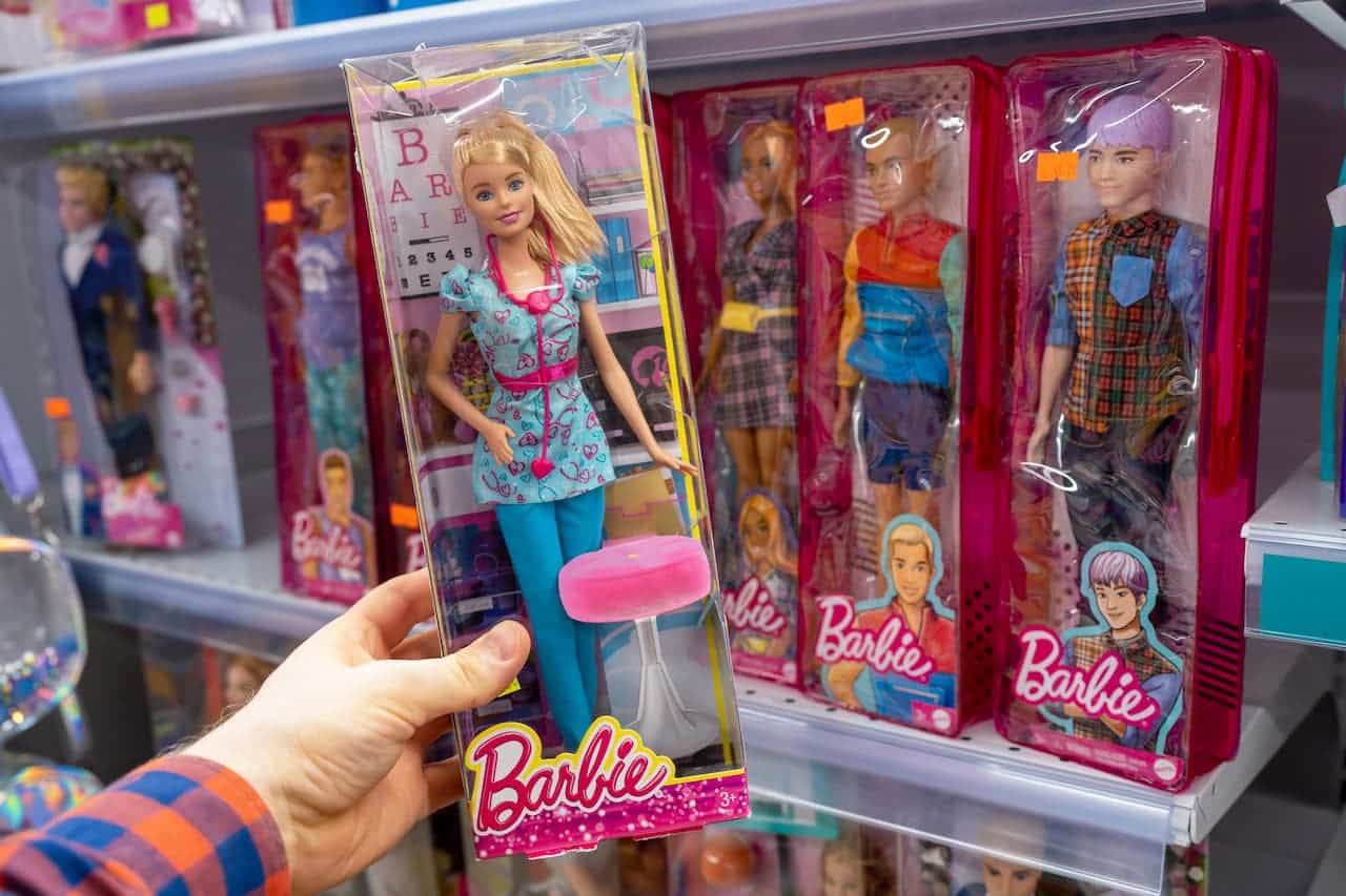 Mão segurando uma embalagem com uma Barbie dentro e bonecas da Barbie ao fundo, numa prateleira