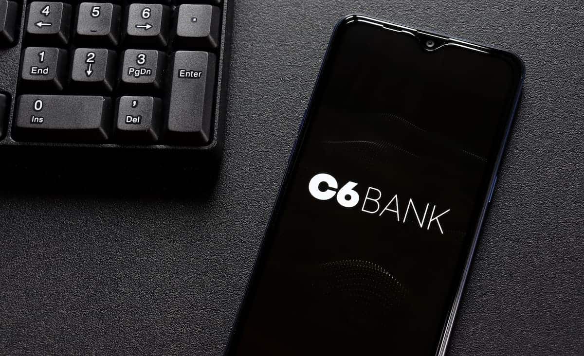 Aplicativo do C6 Bank em celular de cliente sobre mesa, ao lado de teclado de computador
