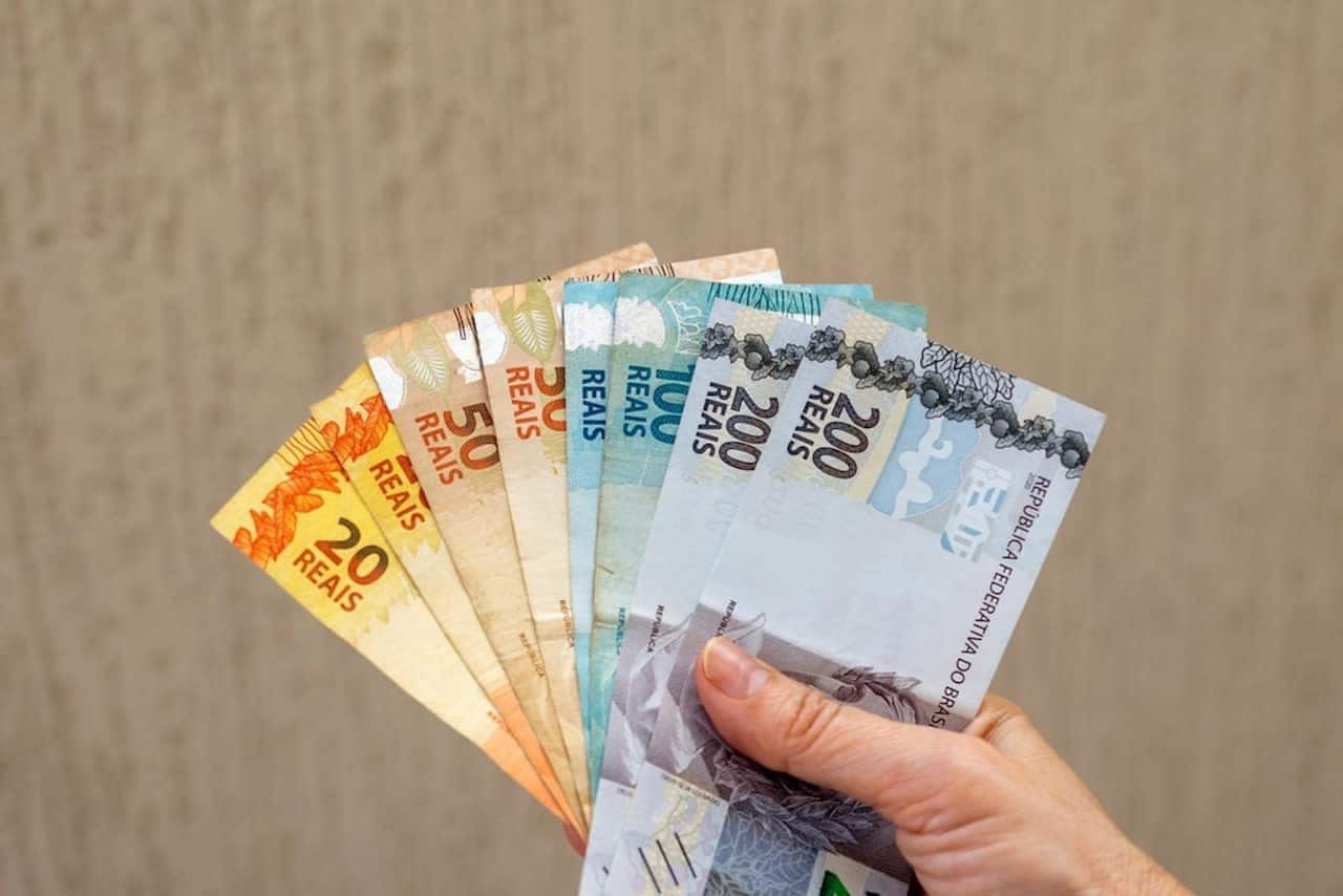 Mão segurando dinheiro, em notas de 20, 50, 100 e 200 reais.