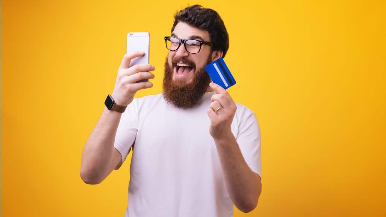 Homem com expressão feliz enquanto olha para celular e segura um cartão de crédito