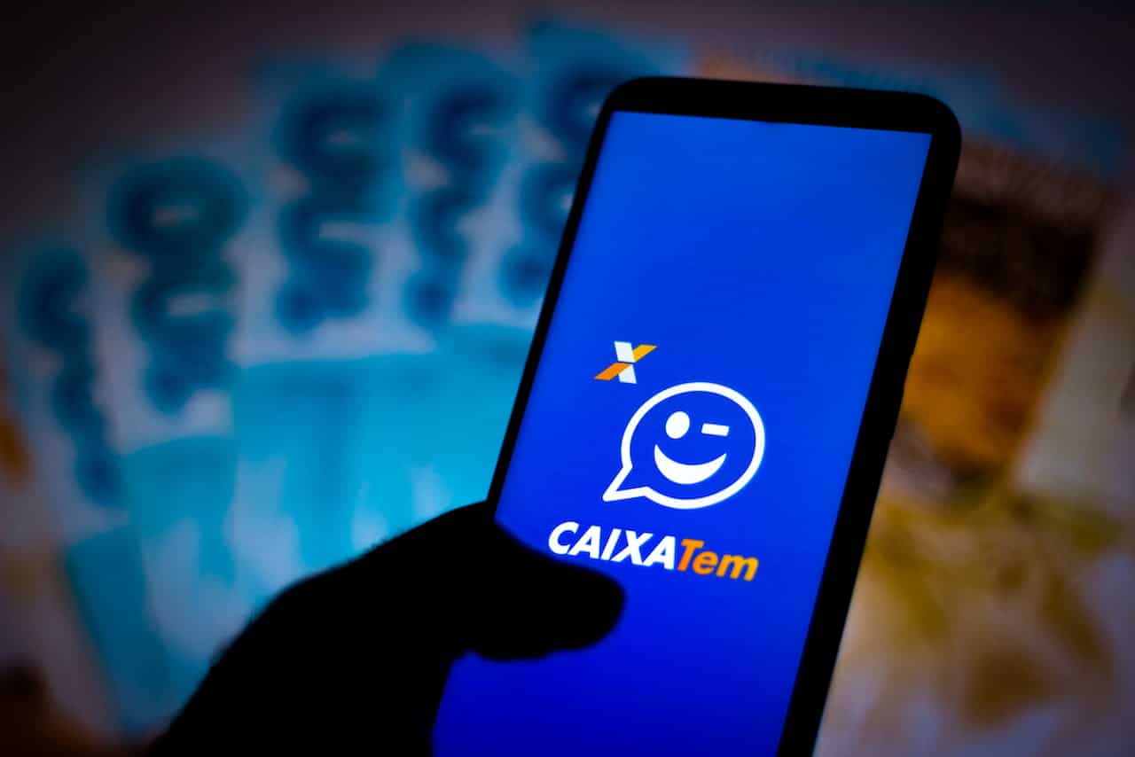 Mão segurando celular com app Caixa Tem aberto. Ao fundo, notas brasileiras embaçadas emprestimo caixa