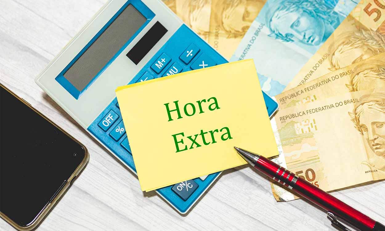Mesa de escritório com calculadora, dinheiro caneta e um post it escrito "hora extra".