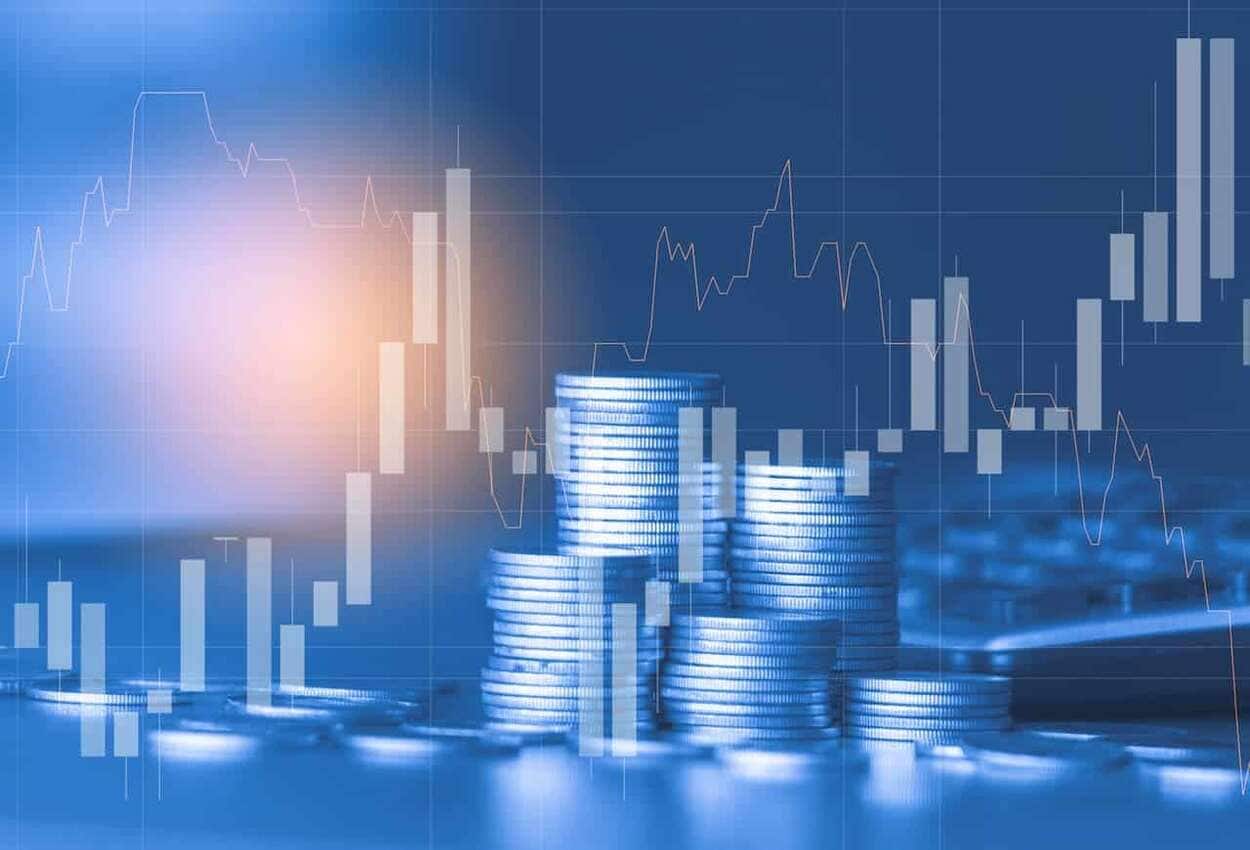 Na imagem, moedas de prata empilhadas em fundo azul com gráficos de investimento.