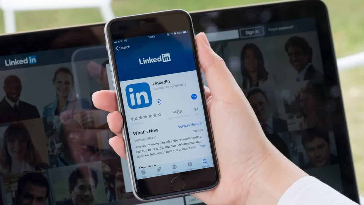 Mão segurando celular aberto no aplicativo do LinkedIn. Ao fundo, um notebook aberto na página do LinkedIn.