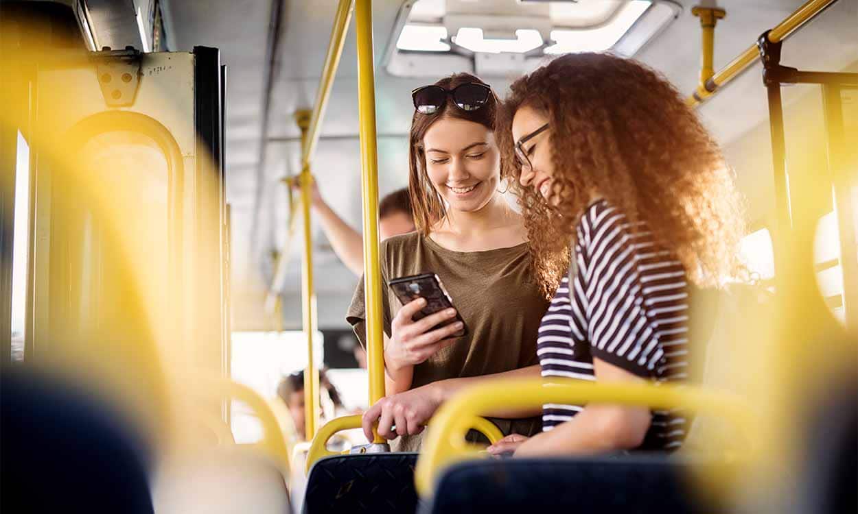 Duas jovens em pé em um ônibus. Elas sorriem e leem notícia em um celular. prazo