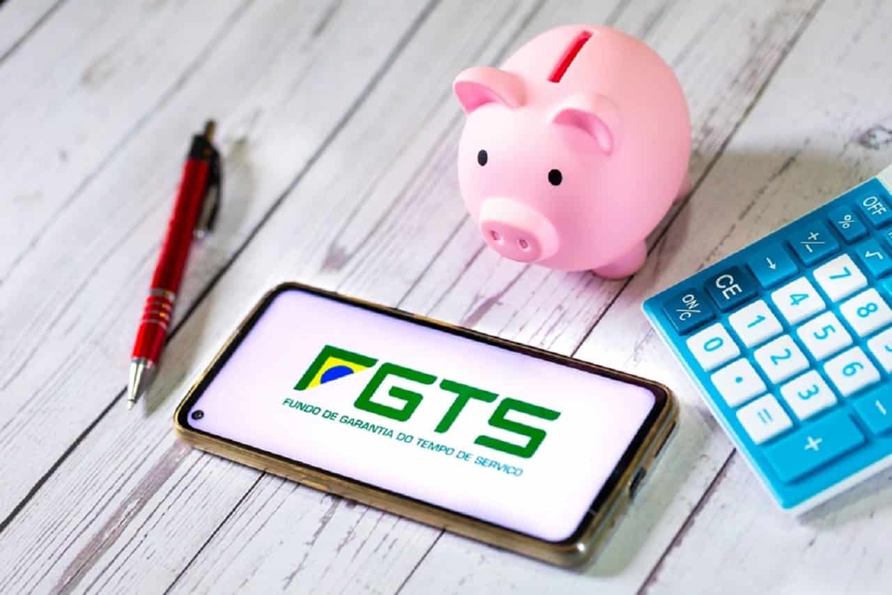 Imagem de um celular com a tela na logo do FGTS. Ao lado, uma calculadora, caneta e um porco em miniatura