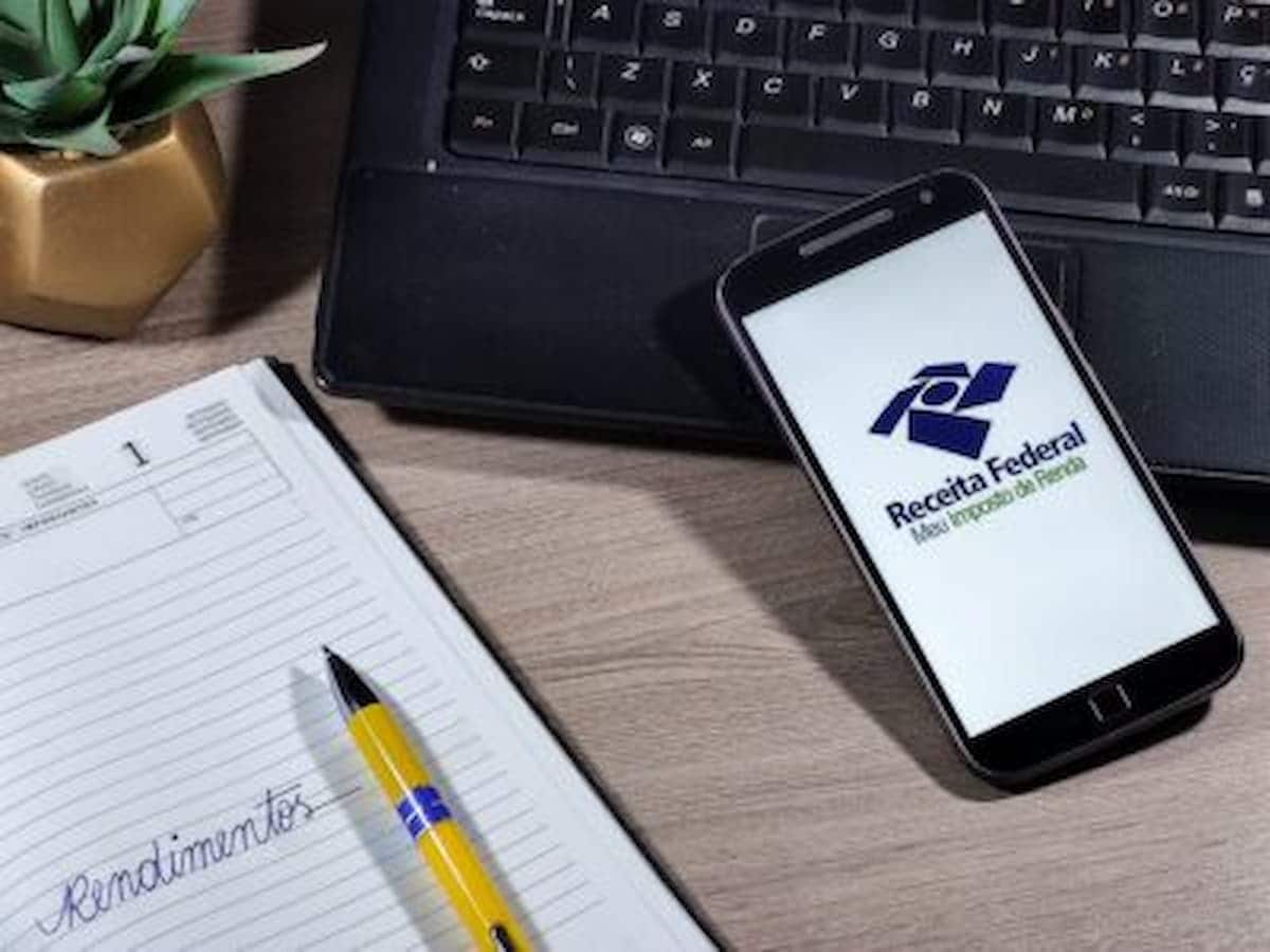 Imagem de um celular com a tela na logo da Receita Federal Imposto de Renda e um caderno ao lado.