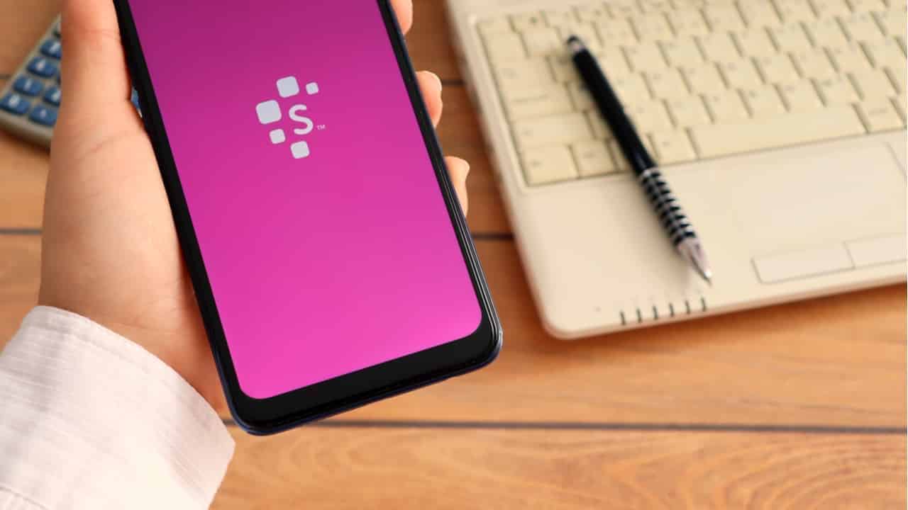 Mão segurando um celular com app Serasa sendo exibido na tela, em segundo plano há um notebook branco, sobre ele há uma caneta preta, ao lado há uma calculadora cinza. Serasa Score
