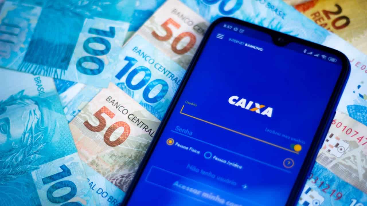 Celular com app Caixa aberto ao lado de notas de 50 e 100 reais.