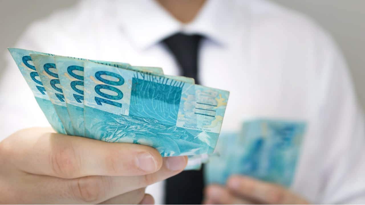 Mão estendendo notas de 100 reais