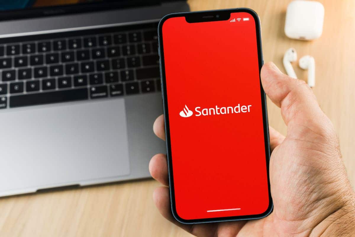 Aplicativo Santander em celular de cliente, seguradoa cima de mesa com notebook e par de fones.