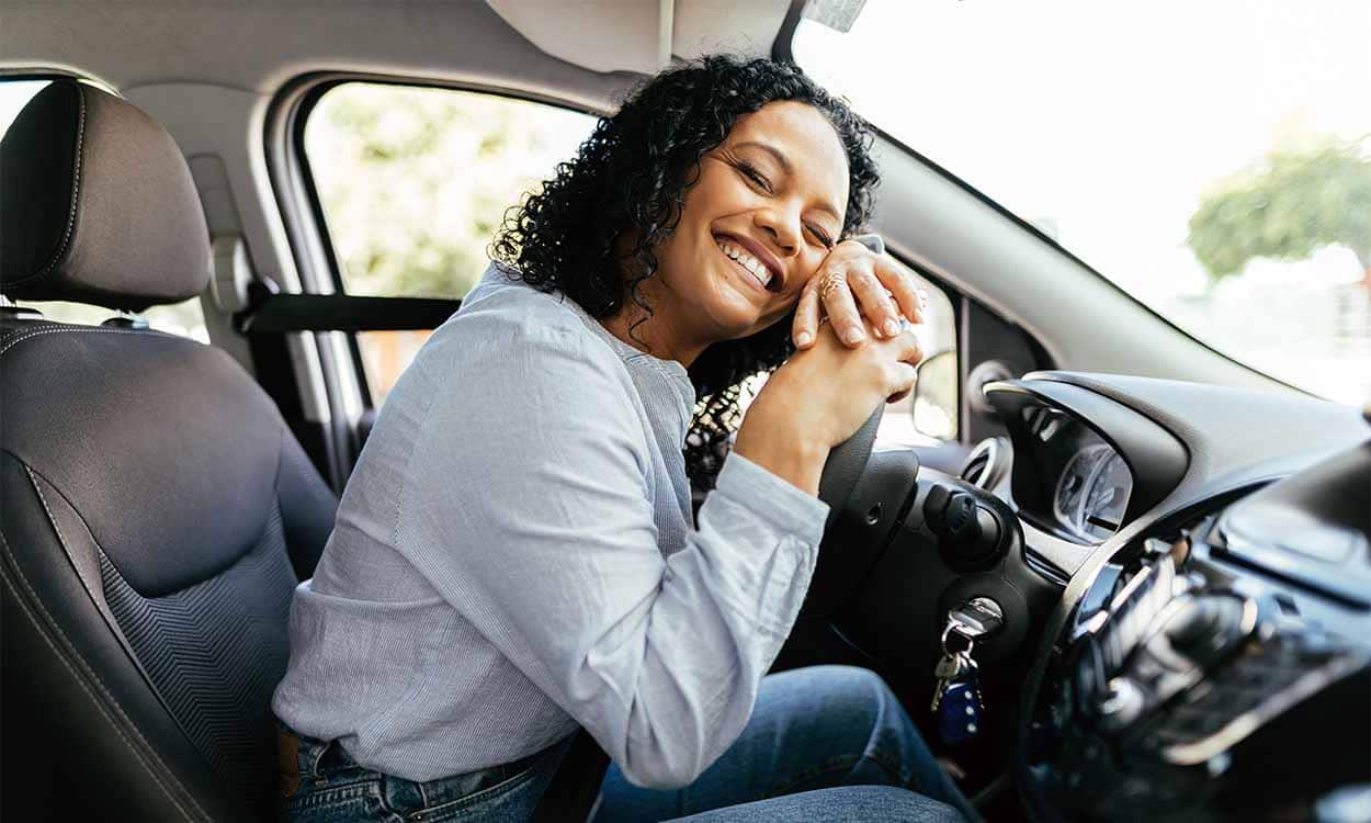 Mulher com expressão de felicidade dentro de um carro, abraçando o volante.