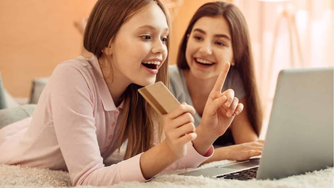 Na imagem, adolescentes na cama com cartão na mão olhando a tela de notebook.
