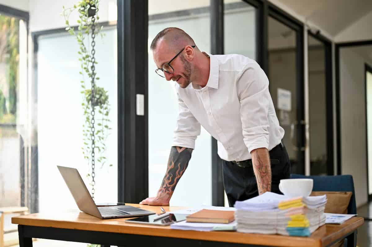 Homem com tatuagem à mostra nos dois braços olha um notebook que está sobre uma mesa