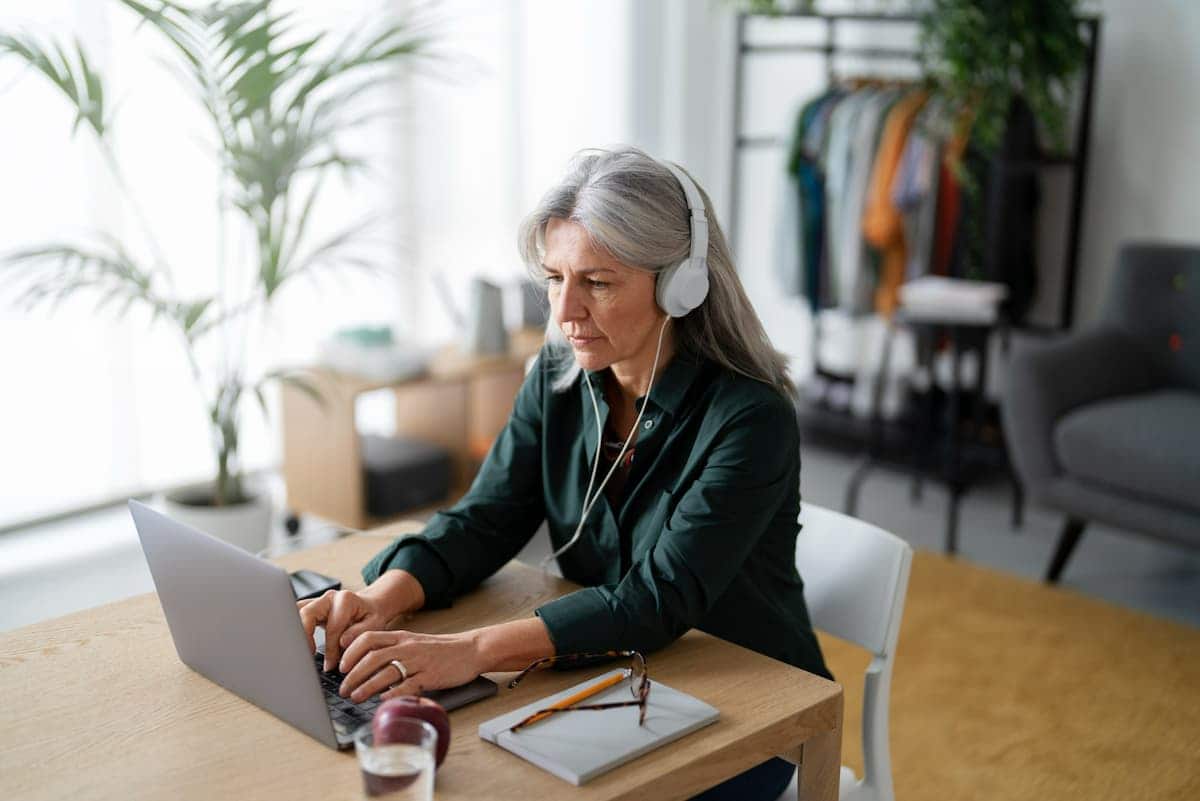 A imagem mostra uma mulher idosa com fones de ouvido, digitando em um notebook. Aparentemente trabalhando.