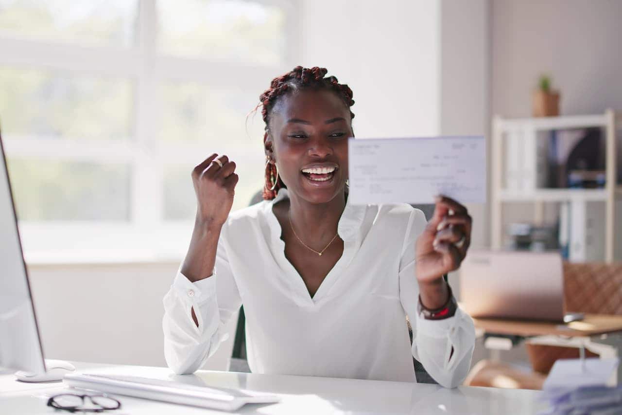 mulher negra com tranças sentada em uma escrivaninha muito feliz com um cheque na mão