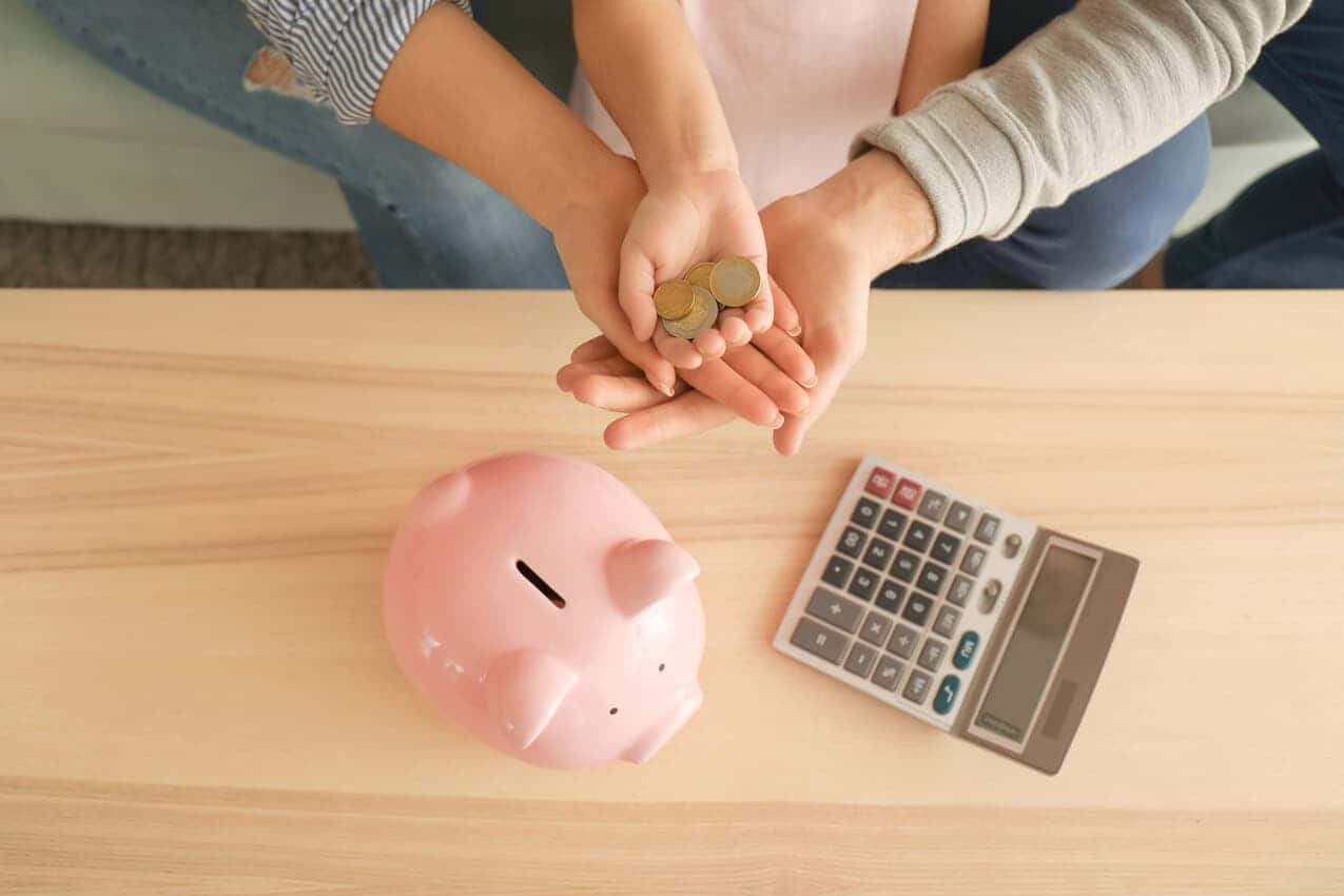 Na imagem, mãos de adulto apoiam uma mão infantil segurando moedas. Sob a mesa abaixo há um cofrinho de porquinho e uma calculadora.