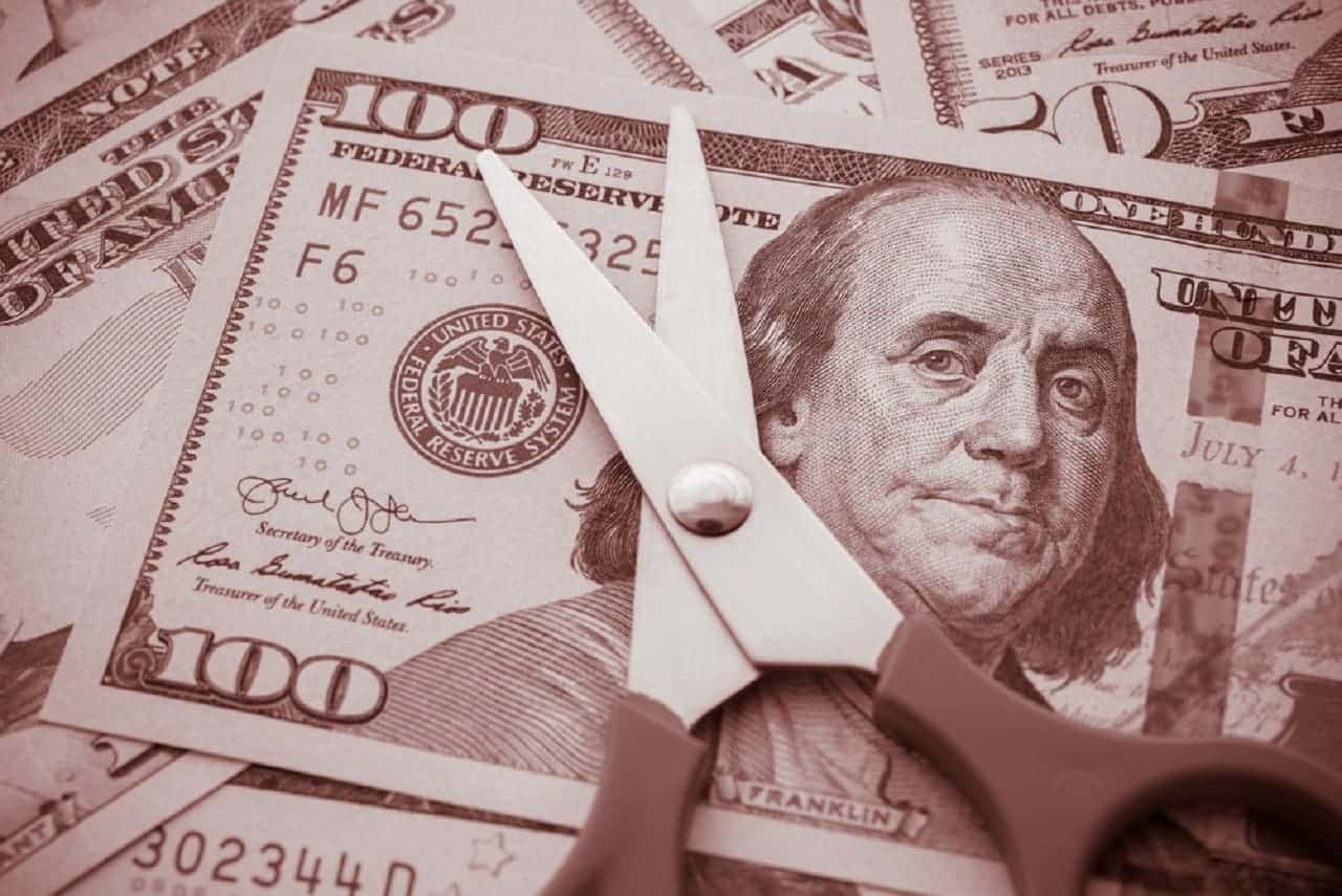 Pequena tesoura posicionada em cima de uma nota de dólar. Várias outras notas compõem a superfície da imagem, que está toda em um tom de sépia.