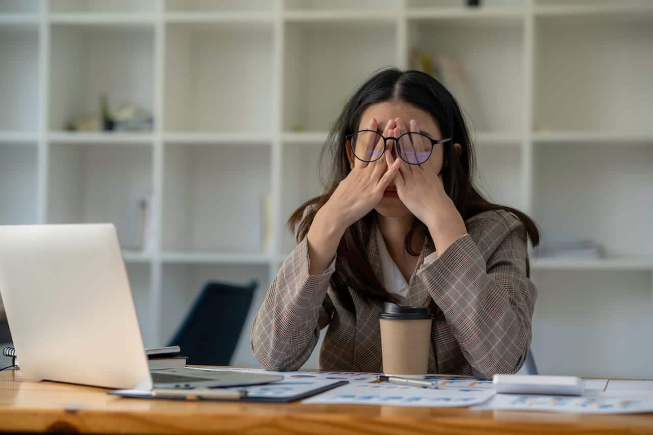 Perto de uma mesa com notebook, papéis e copo, mulher cobre os olhos com as mãos