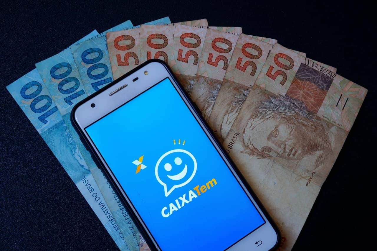 Celular com app Caixa Tem aberto e em cima de várias notas de 50 e 100 reais.
