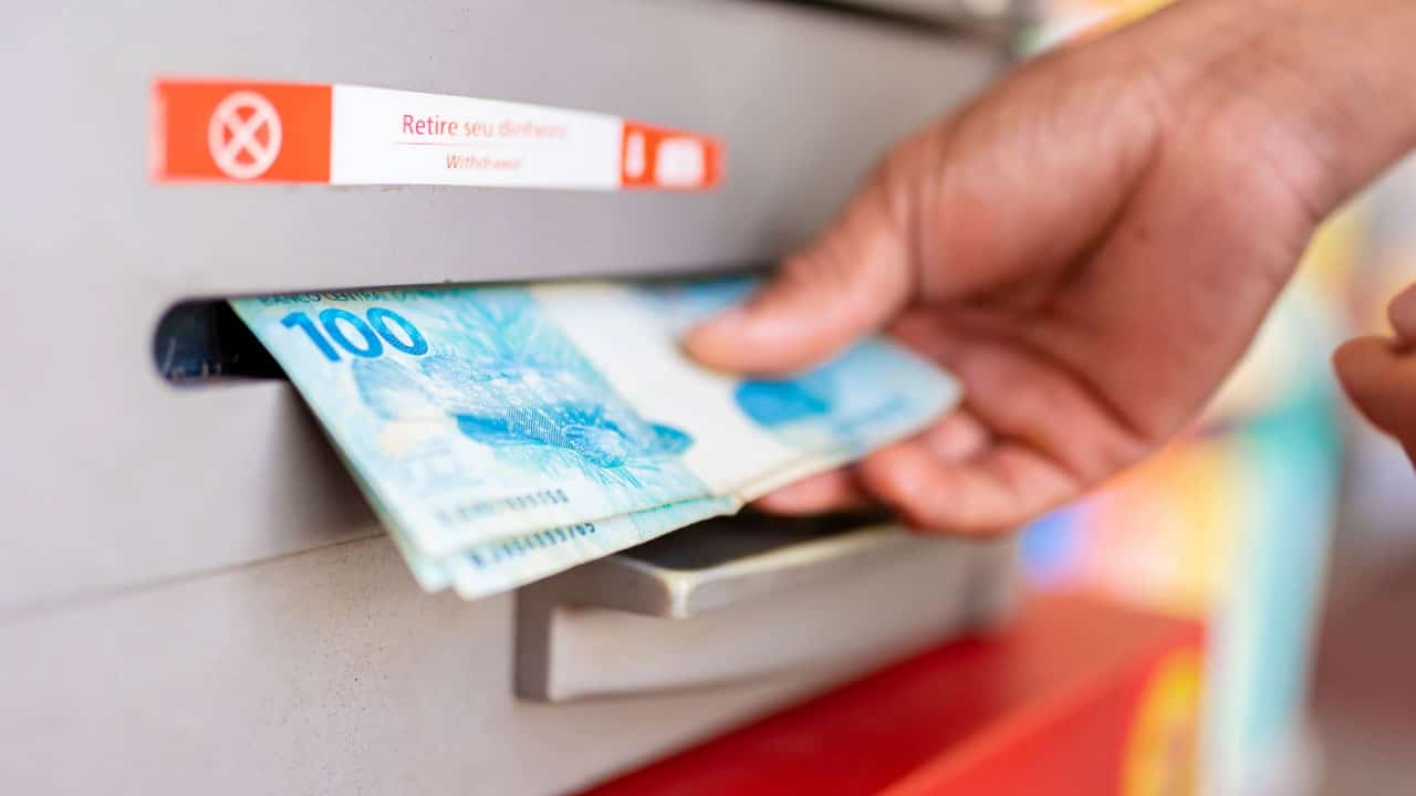 Mão retirando notas de 100 reais da boca do caixa eletrônico