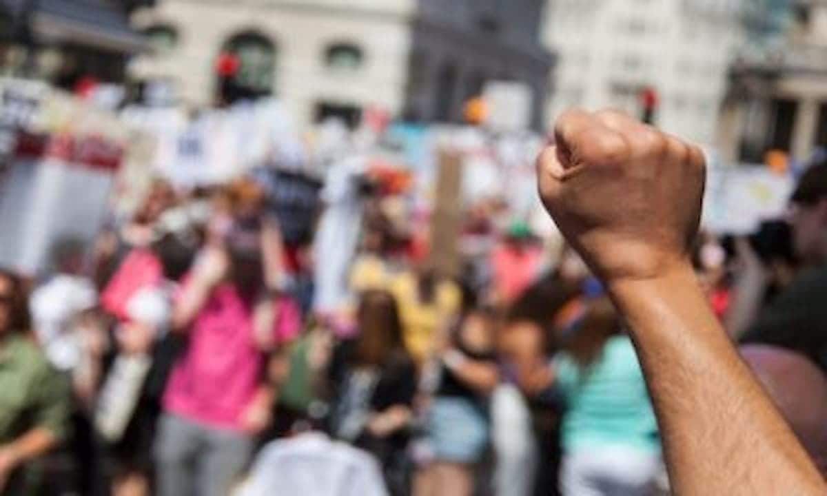 Pessoas aglomeradas em um protesto, com destaque ao braço de uma pessoa negra que está com a mão fechada simbolizando resistência