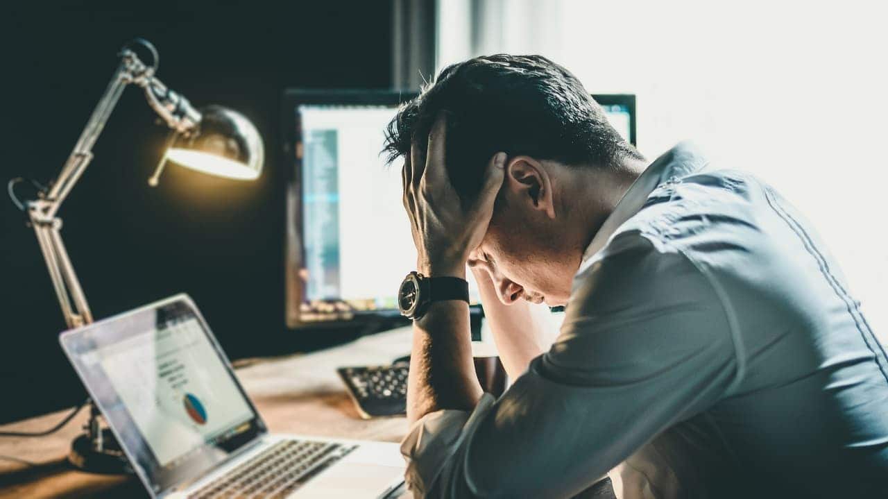 Homem com cabeça apoiada nas mãos em uma mesa com computador.