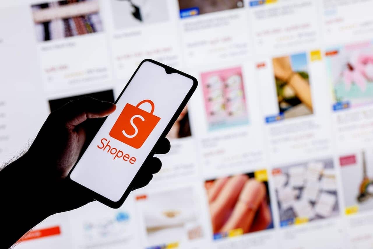Mão segurando celular com logo Shopee. Ao fundo, tela com diversos anúncios na plataforma