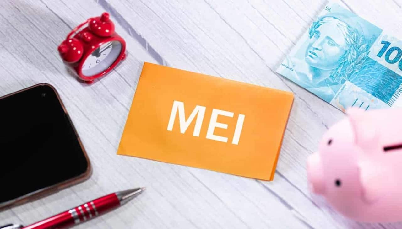 A imagem mostra um post-it laranja escrito "MEI" e ao lado um cofre de moedas em formato de porquinhos, uma nota de cem reais, um despertador, bloco e caneta.