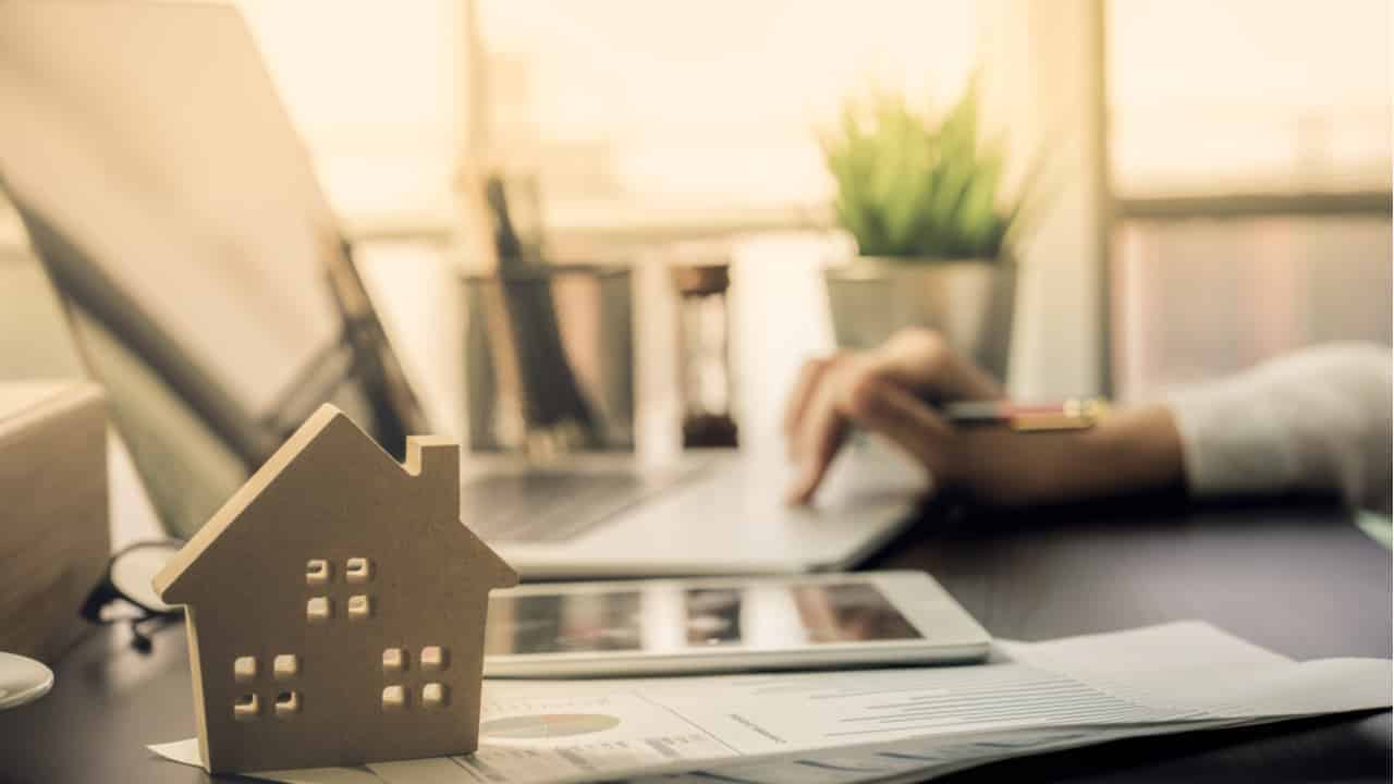 Na imagem, pessoa digita no laptop e na mesa há um enfeite representando uma casa.