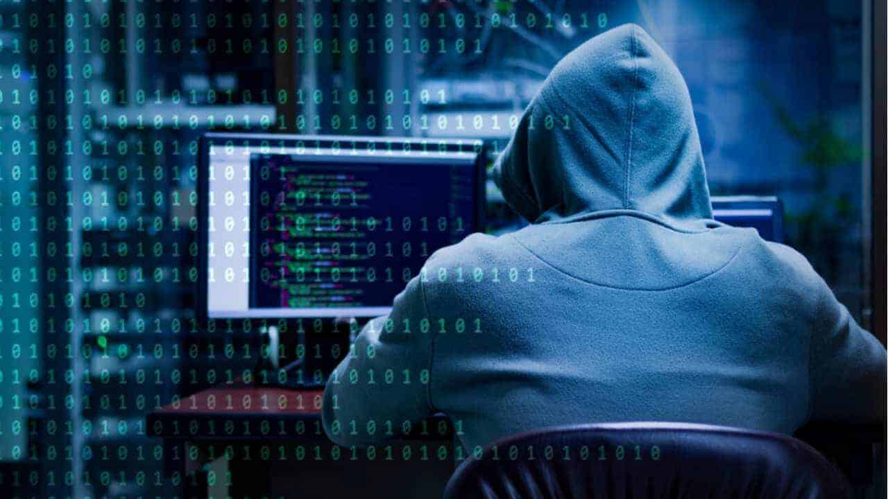 Pessoa com capuz, de costas, em frente a um computador em ambiente escuro