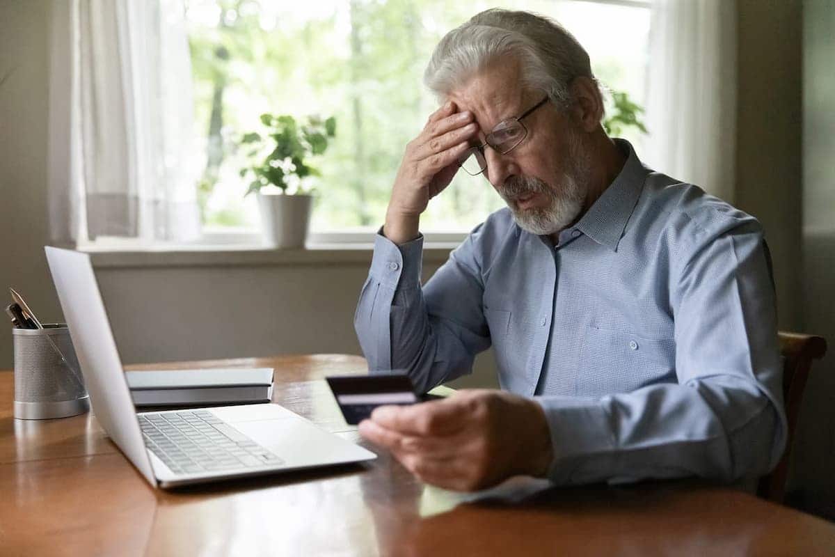 Idoso em frente ao notebook, segurando um cartão de crédito, com expressão de preocupação.