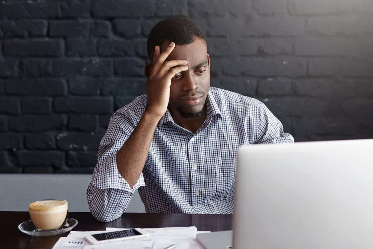 A imagem mostra um jovem, com o notebook aberto, e com a mão na cabeça, aparentemente preocupado.