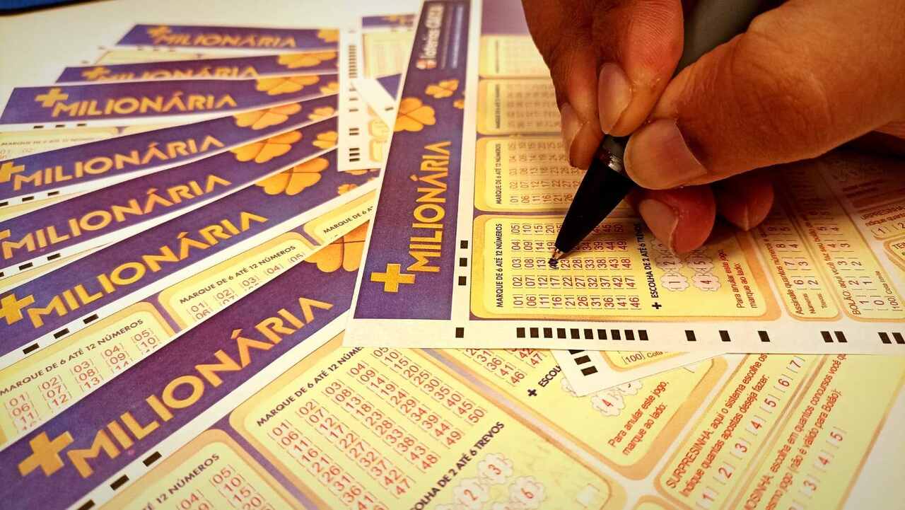 Diversos volantes da loteria +Milionária e uma pessoa preenchendo uma cartela, utilizando uma caneta preta