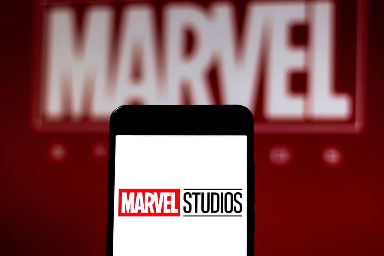 Celular com tela branca e logomarca da Marvel Studios na frente de um painel vermelho com a logo da Marvel