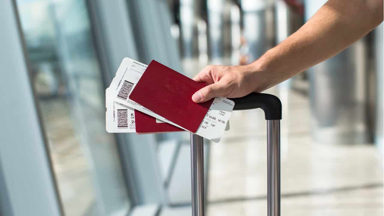 Imagem com foco na mão de um rapaz segurando 2 passagens de avião, com o braço apoio na alça da mala para viajar