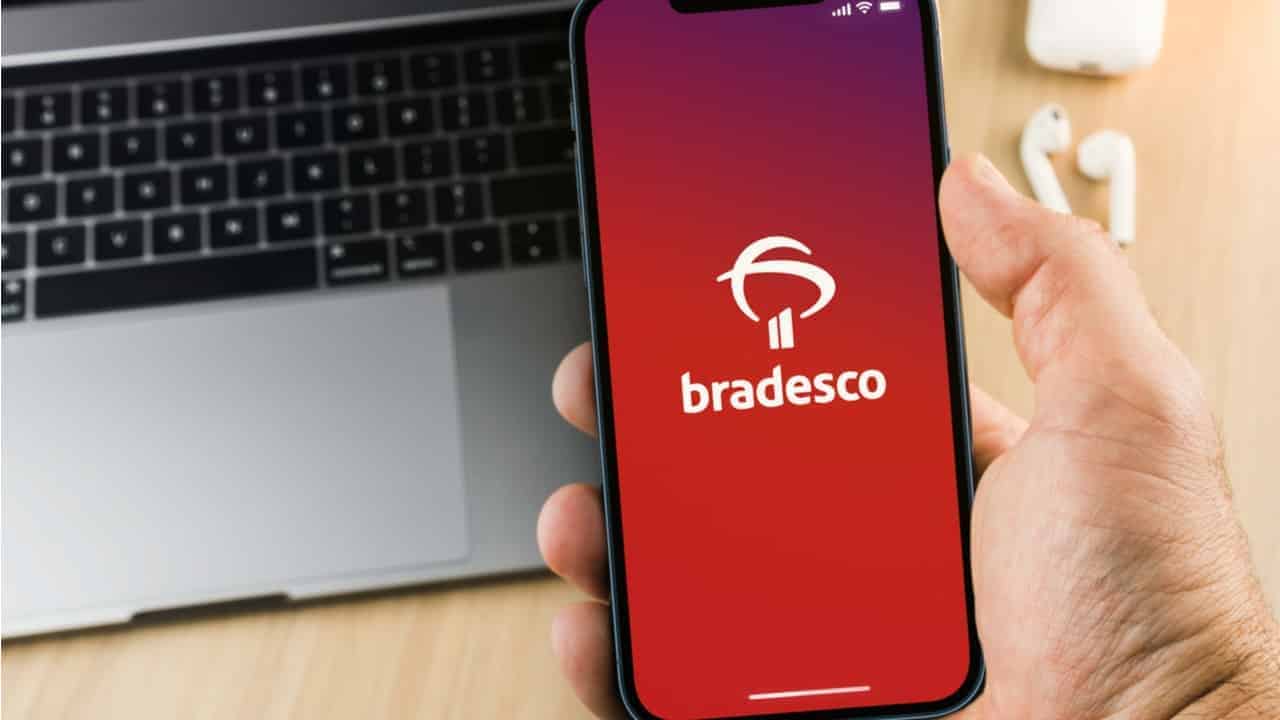 Uma mão segurando um celular utilizando o aplicativo do Bradesco, no fundo aparece um notebook
