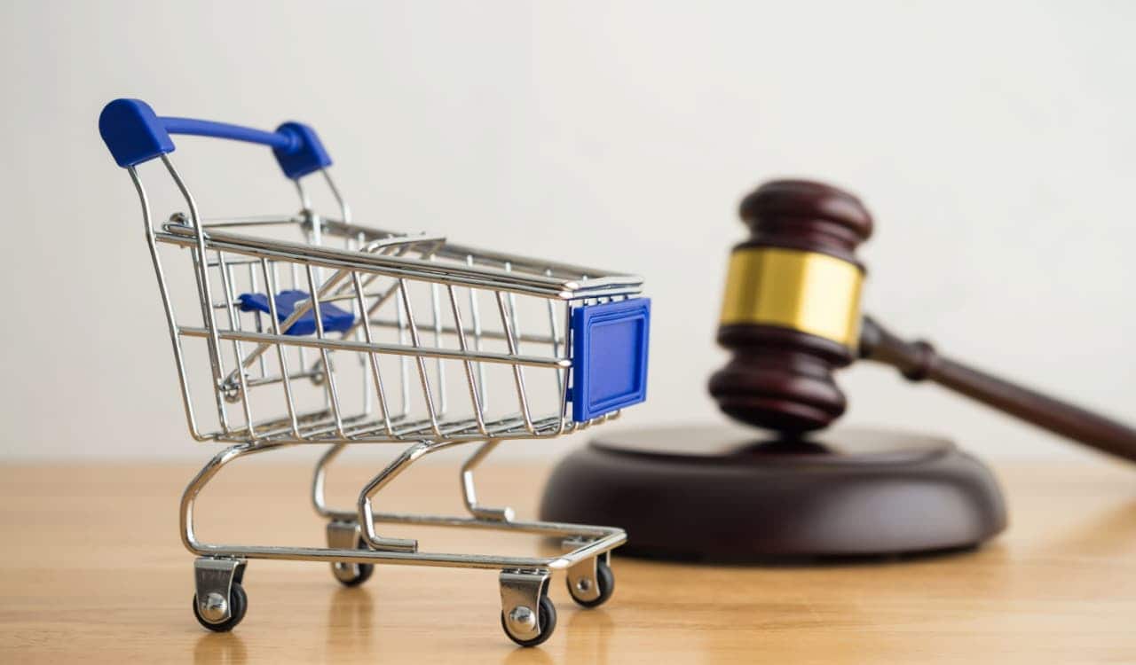 Carrinho de compras pequeno em cima de uma mesa com um martelo de madeira ao fundo em cima de um disco de madeira marrom, representando a justiça e os direitos do consumidor