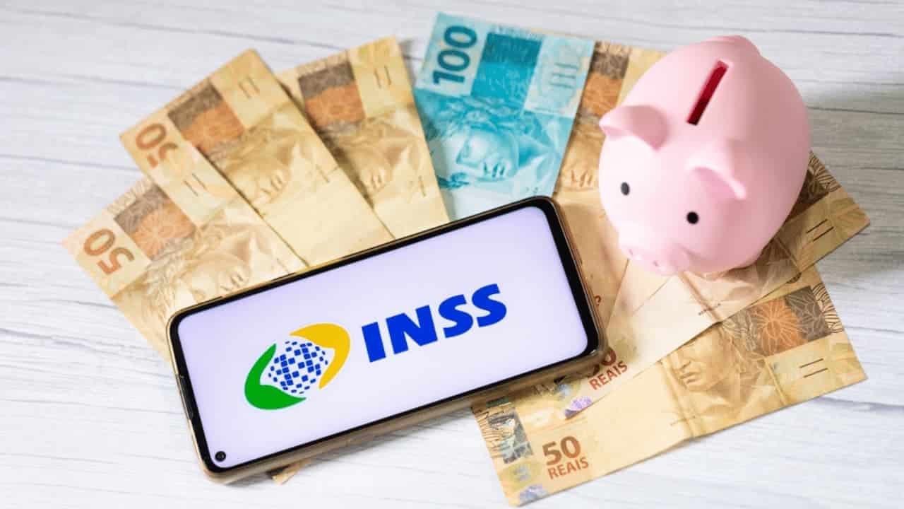 Um celular com a logo do INSS e um cofrinho em formato de porco sobre várias notas de 100 e 50 reais.