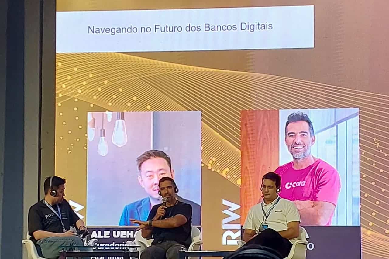 Telão ao fundo escrito “Navegando no Futuro dos Bancos Digitais”. À frente, três homens sentados em conversa.