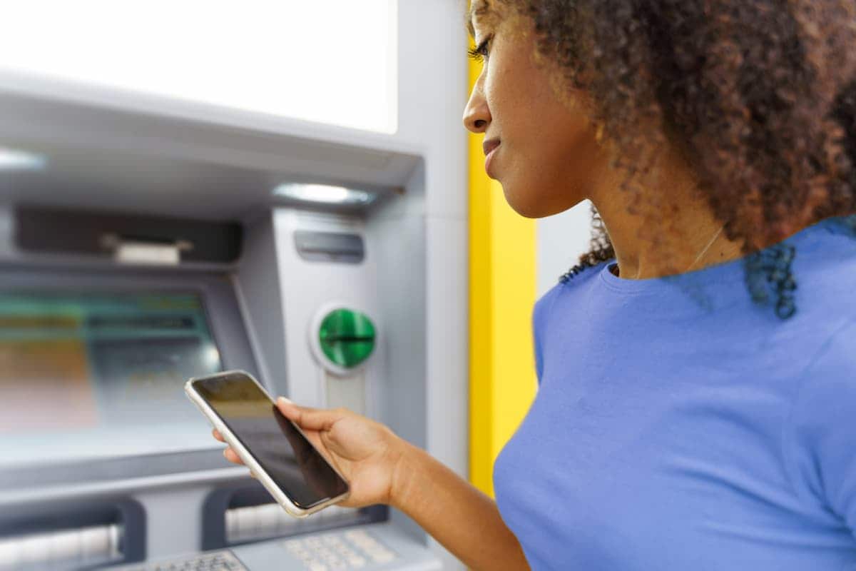 Imagem de mulher mexendo em aplicativo de banco, em frente a caixa eletronico
