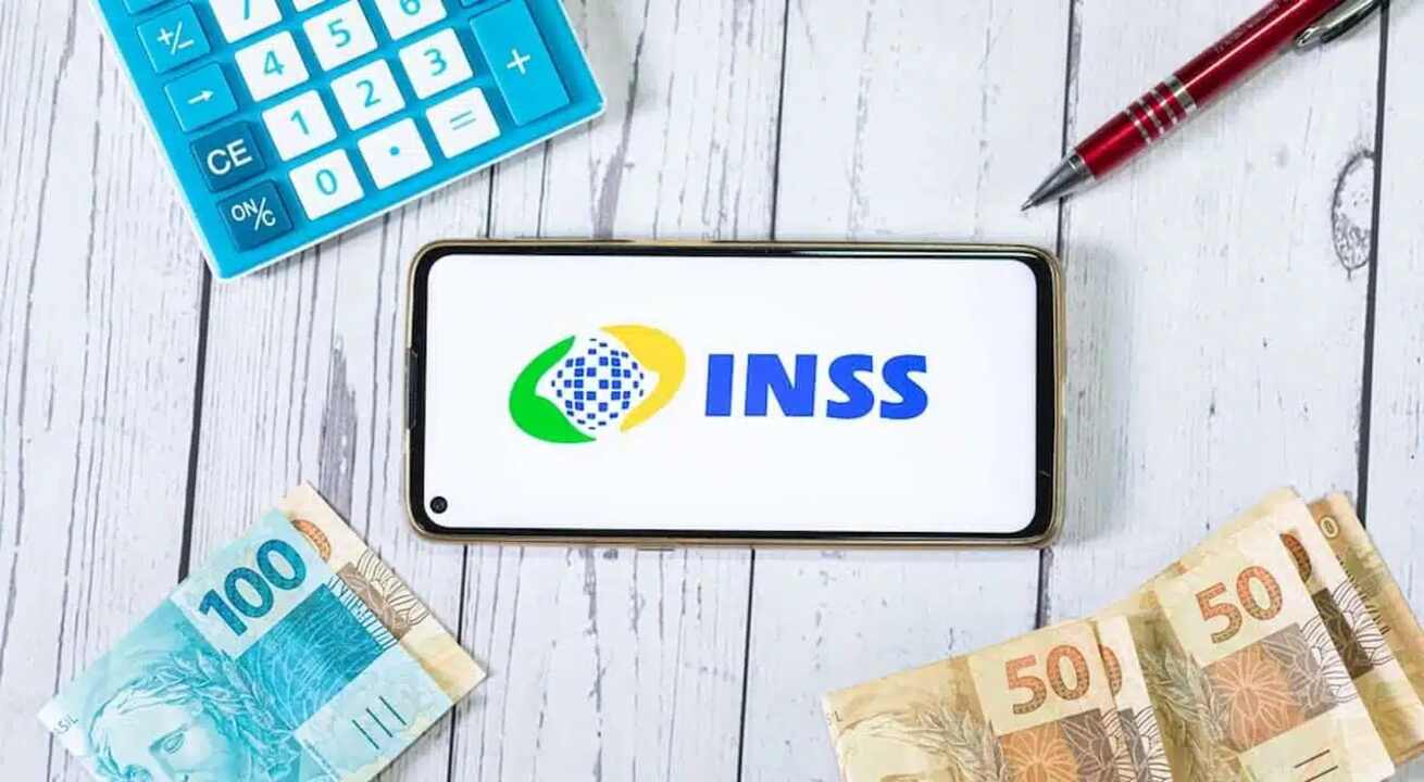 Celular com logo INSS e calculadora, caneta e cédulas brasileiras ao redor.