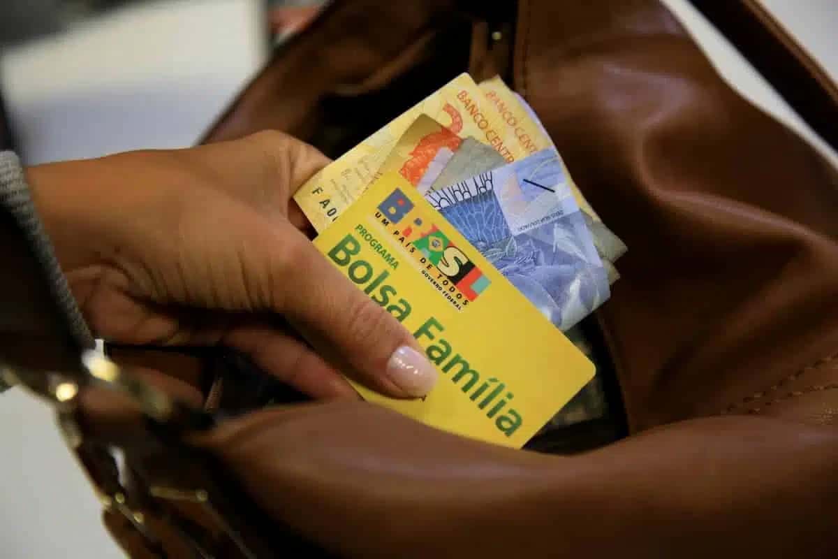 mão de mulher negra segurando cartão do Bolsa Família e cédulas de real e colocando esses itens dentro de uma bolsa marrom