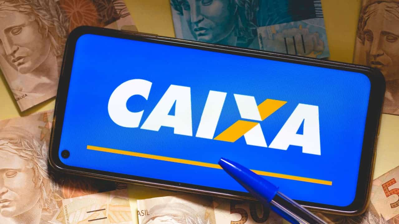 Celular com logo Caixa sobre algumas notas de dinheiro na mesa.