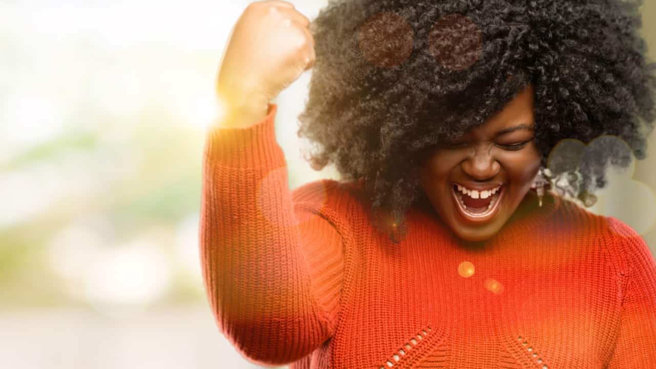 Mulher negra, de cabelos cacheados e pretos, comemorando em pose de vitória, sorrindo
