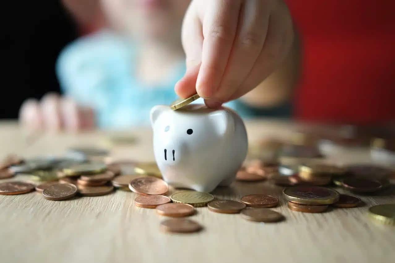 Mão de uma criança colocando moeda em cofre de porquinho. Moedas espalhadas na superfície.