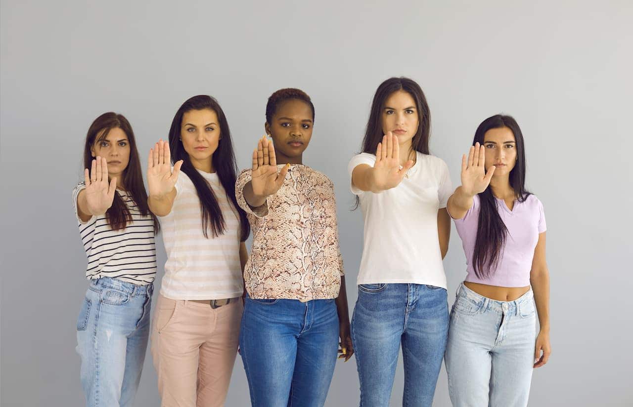 cinco mulheres lado a lado, de diferentes etnias, com uma mão à frente, pedindo o fim da violência contra mulheres