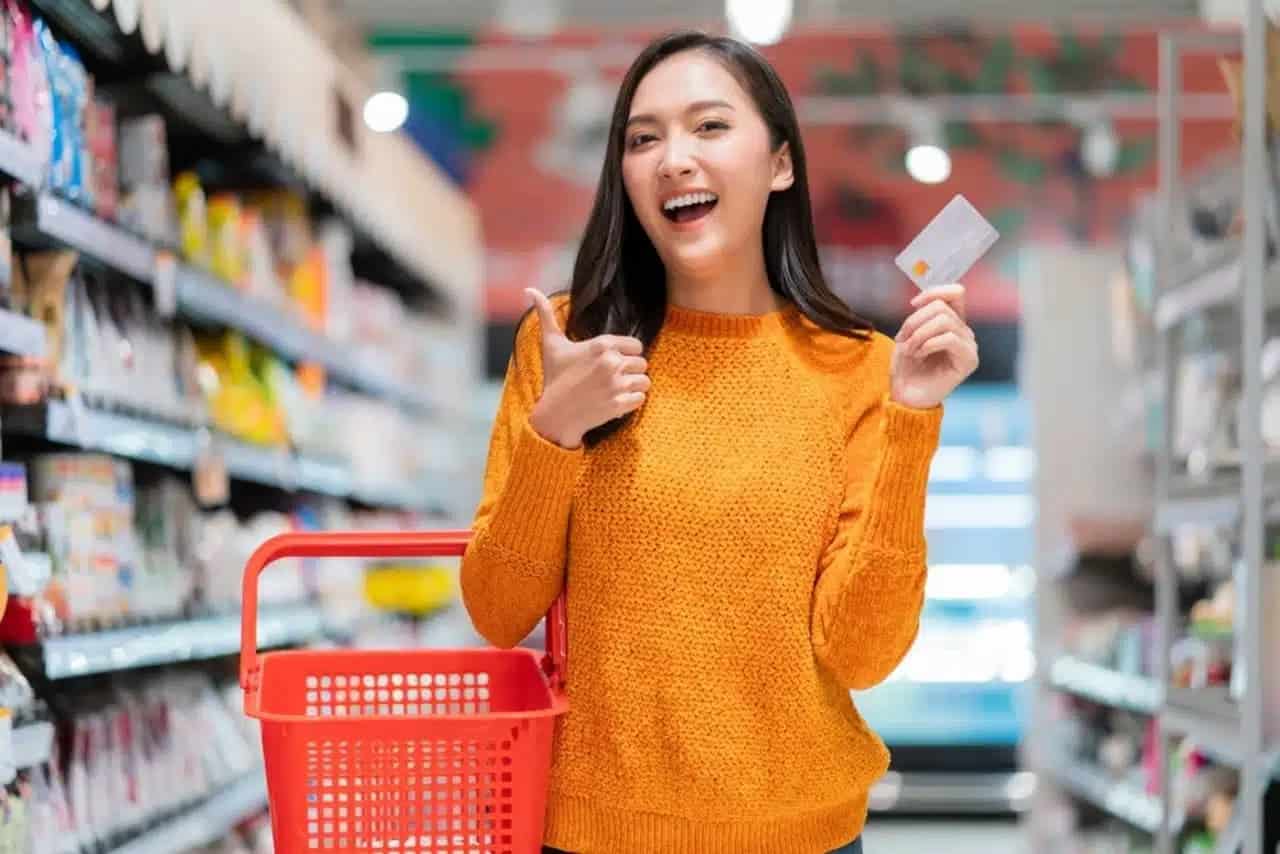 Mulher sorridente no interior de um supermecado segurando uma cesta de compras e um cartão de crédito.